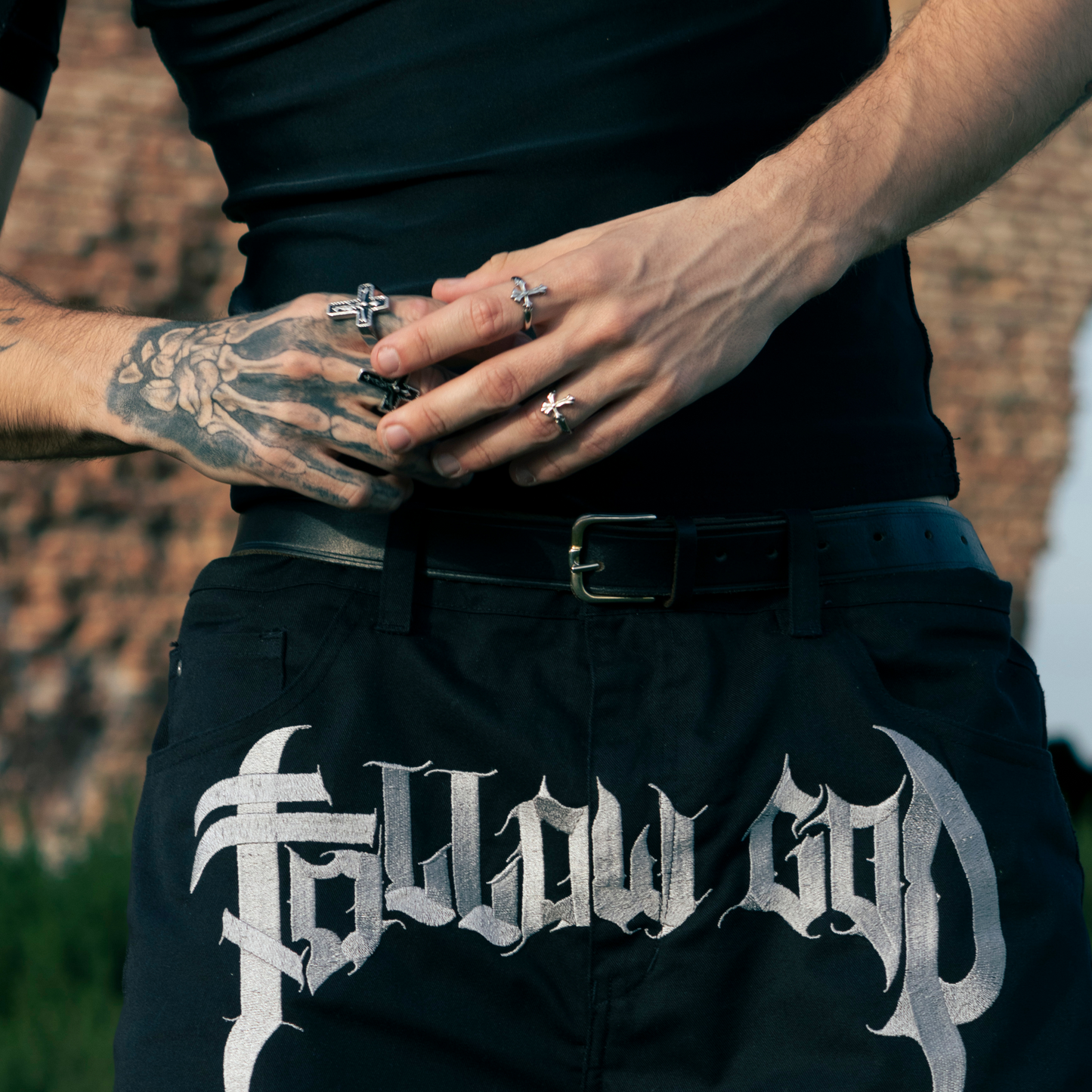 Mano tatuada luciendo múltiples anillos de acero inoxidable 'Follow Ring Big Cross' y 'Follow Ring Lil Cross', combinados con los pantalones 'Salmo Pant' de la marca Follow God con el logo visible