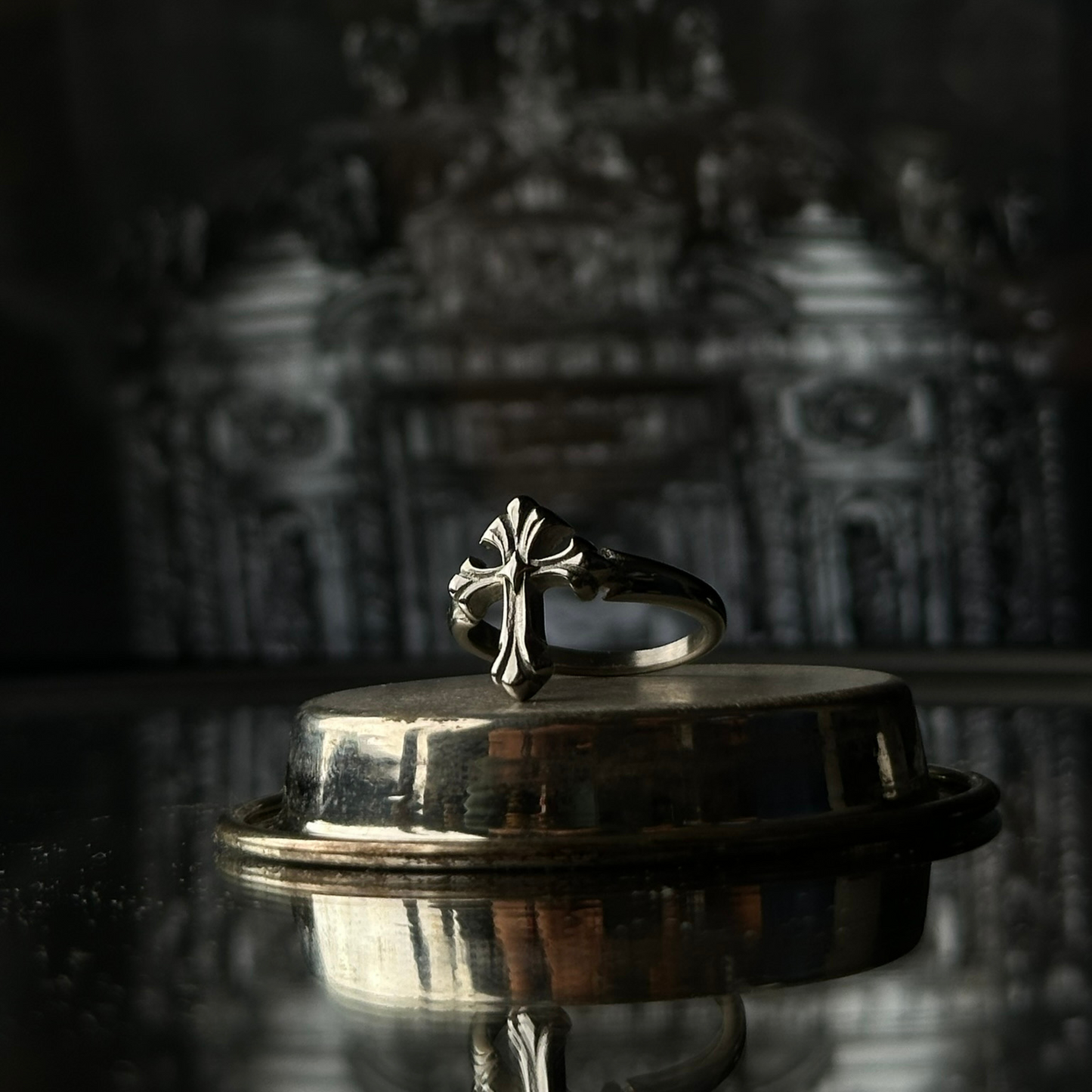 Anillo de acero inoxidable 'Follow Ring Lil Cross' con diseño de cruz en relieve, de la marca Follow God, exhibido sobre una superficie metálica con un fondo arquitectónico