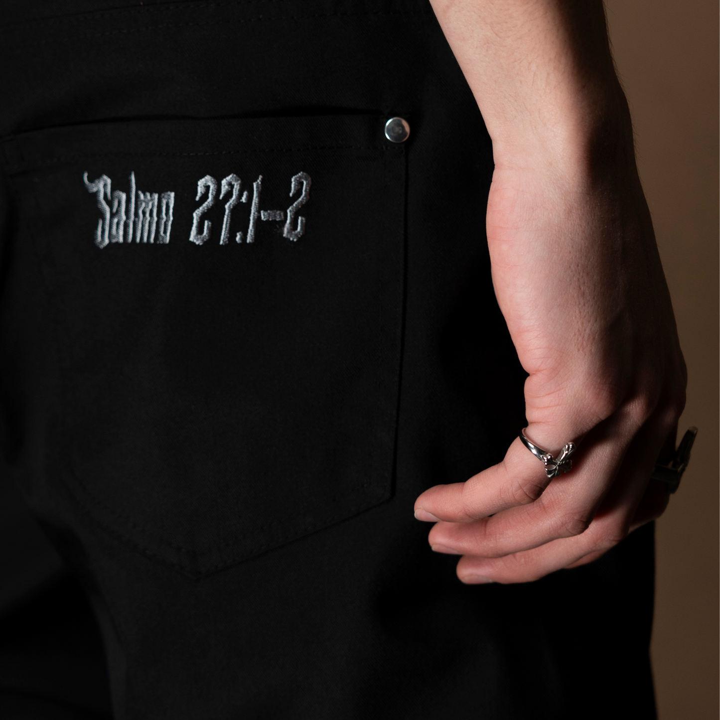 Mano luciendo los anillos de acero inoxidable 'Follow Ring Lil Cross' y 'Follow Ring Big Cross', junto a la parte trasera de los pantalones 'Salmo Pant' con la inscripción del Salmo 27:1-2, de la marca Follow God