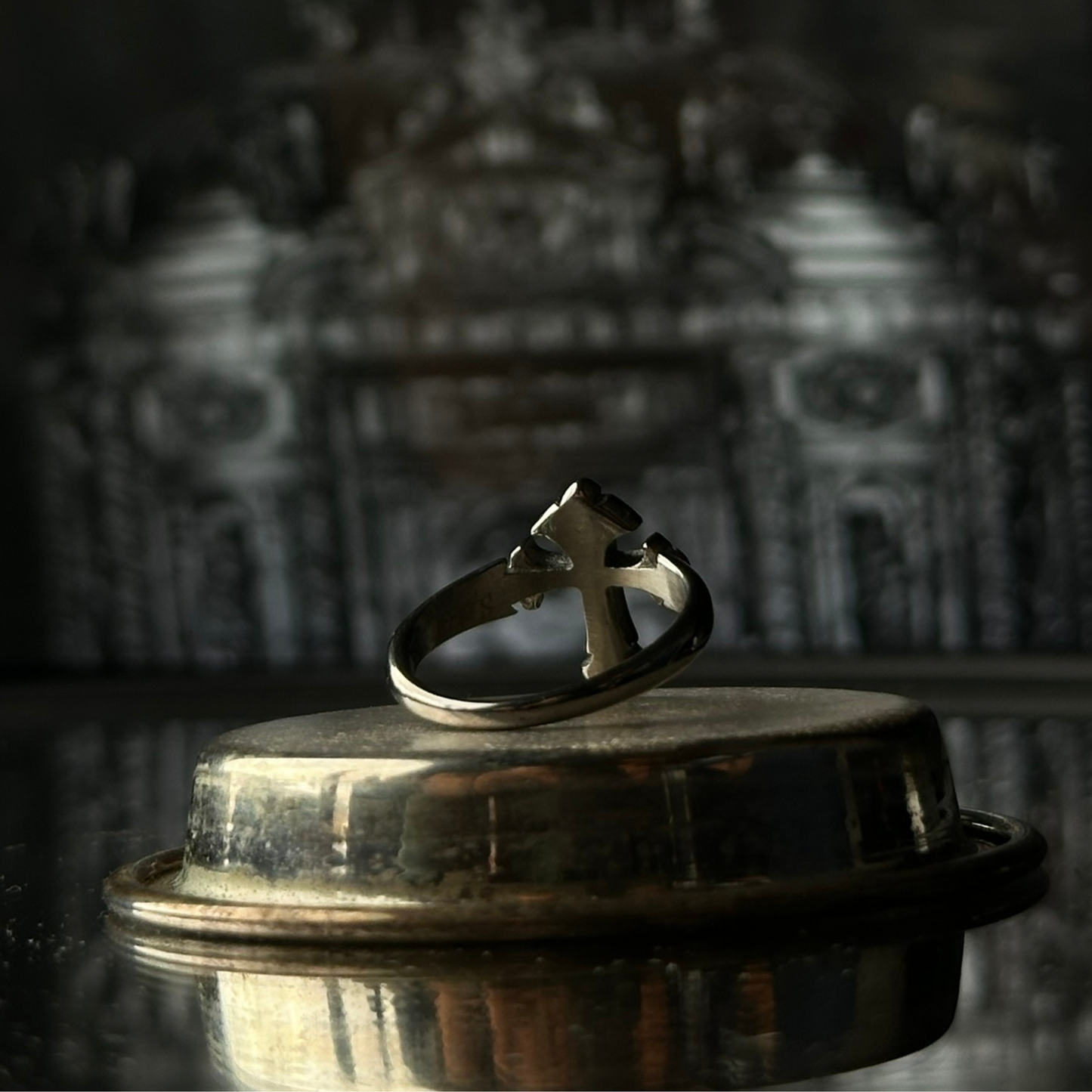 Vista trasera del anillo de acero inoxidable 'Follow Ring Lil Cross' con diseño de cruz en relieve, de la marca Follow God, exhibido sobre una superficie metálica con fondo arquitectónico