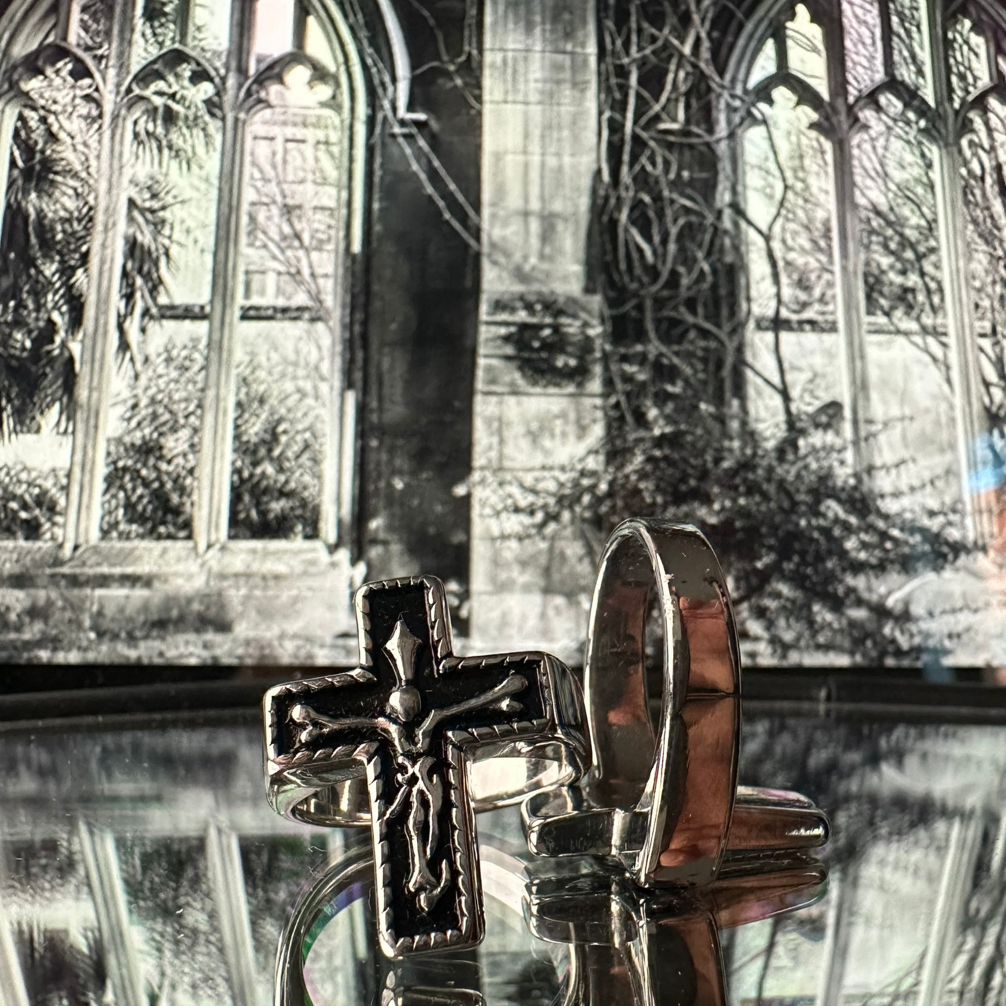 Dos vistas del anillo de acero inoxidable tipo sello 'Follow Ring Big Cross', de la marca Follow God, frente a un fondo arquitectónico de ventanas góticas