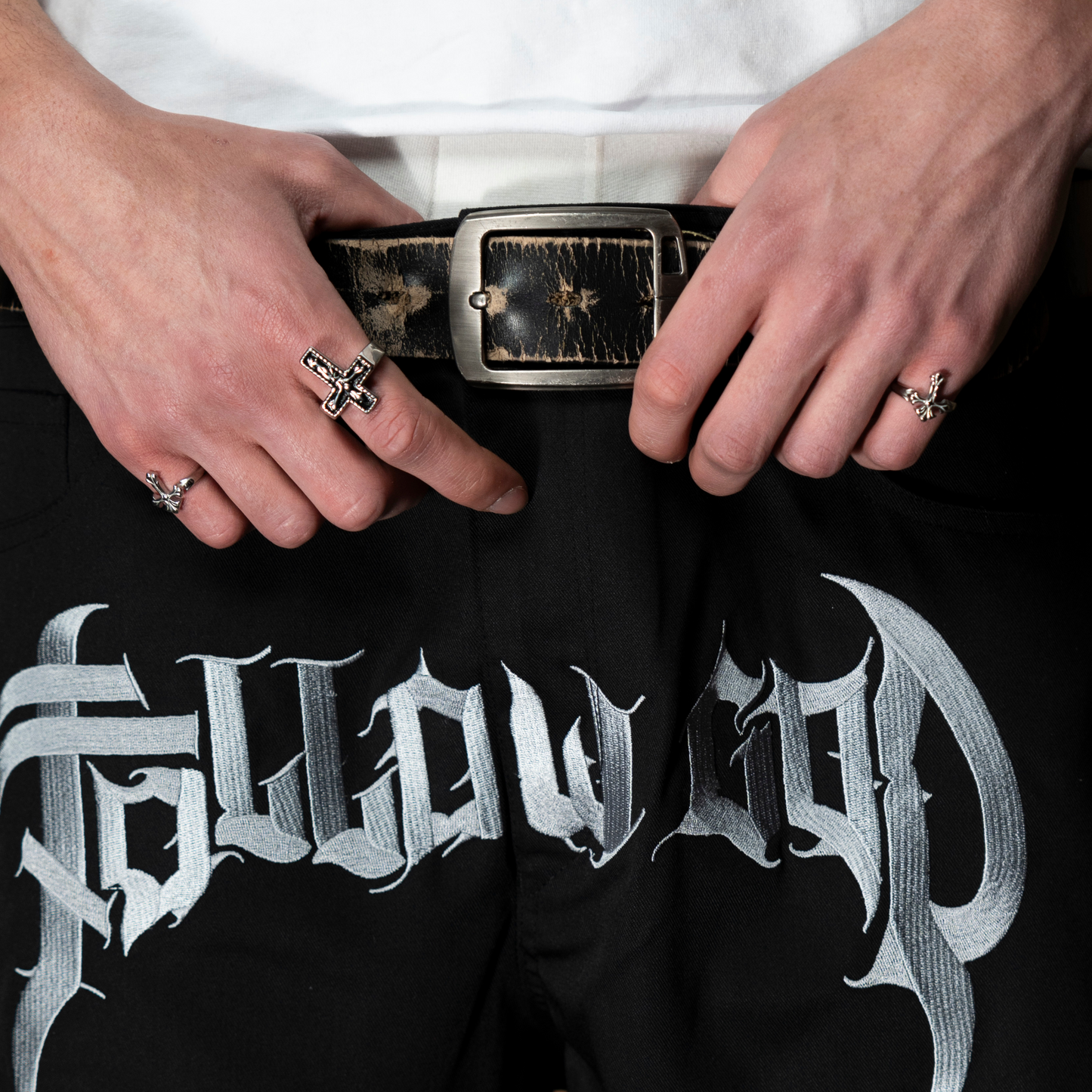 Manos luciendo los anillos de acero inoxidable 'Follow Ring Lil Cross' y 'Follow Ring Big Cross', combinados con los pantalones 'Salmo Pant' de la marca Follow God con el logo visible en la parte frontal